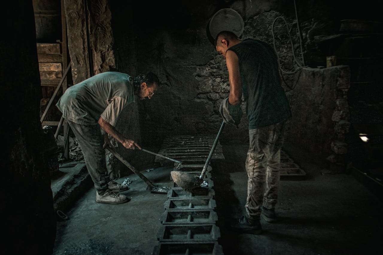Two men working with molten metal in a dimly lit foundry setting.